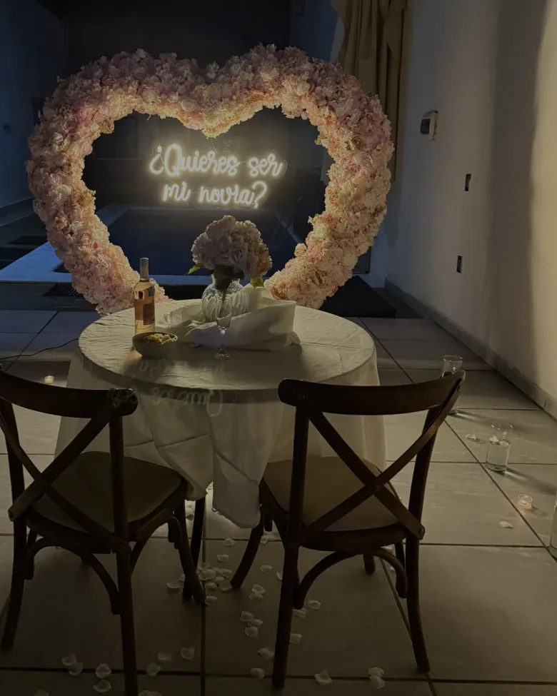 La mesa para dos con mantel, vino, flores y copas, frente al corazón de flores con el neón «¿Quieres ser mi novia?»