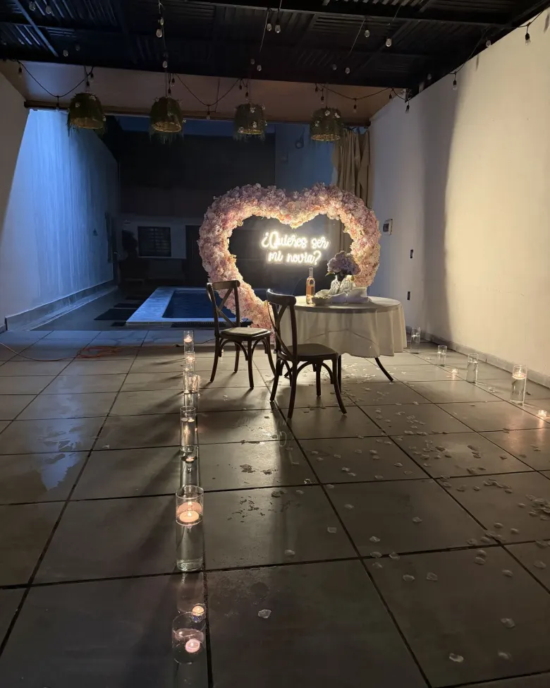 La terraza de Terraza Plutarco de noche, lista para una pedida: el corazón de flores con el neón «¿Quieres ser mi novia?» encendido, la mesa para dos y la alberca iluminada al fondo, con un camino de velas