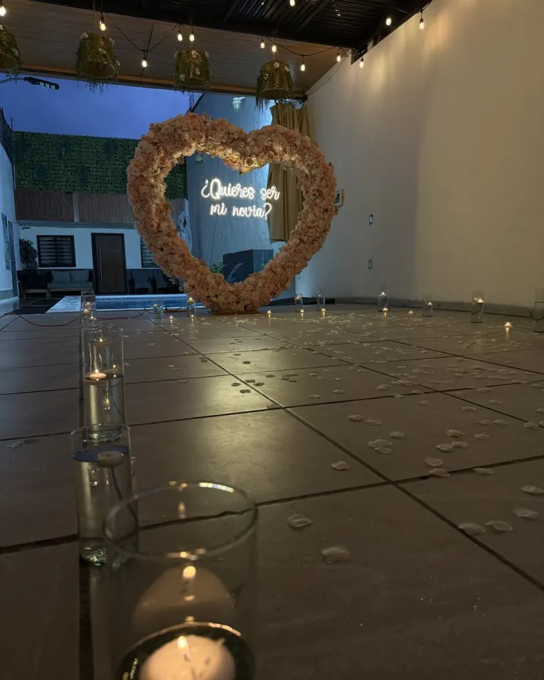 Una vela flotante encendida en primer plano y, al fondo, el corazón de flores con el neón junto a la alberca
