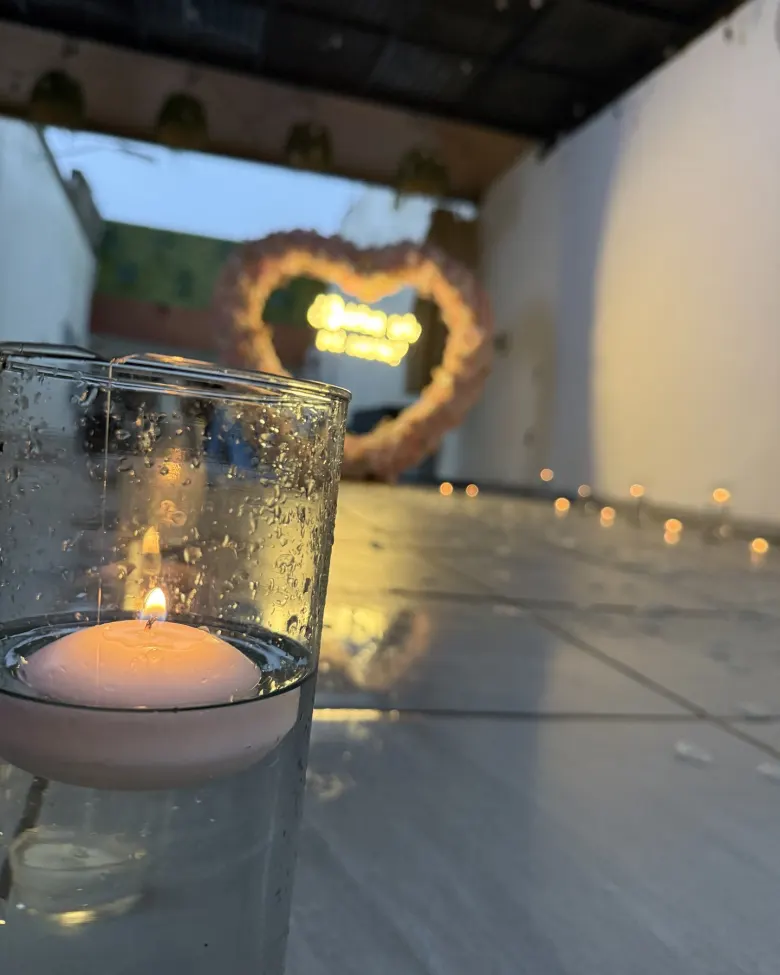 Una vela flotante encendida sobre el piso de la terraza y el corazón de flores iluminado, desenfocado al fondo